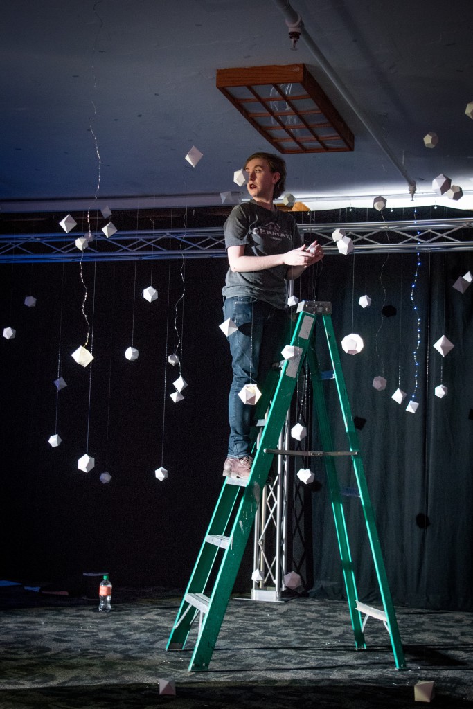 One of the students on a ladder installing hanging paper art on the ceiling