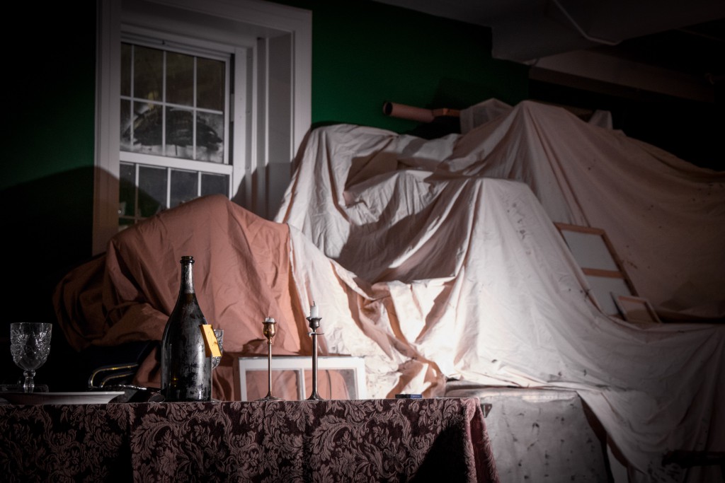 bedsheets cover furniture behind a table with candlesticks and a bottle