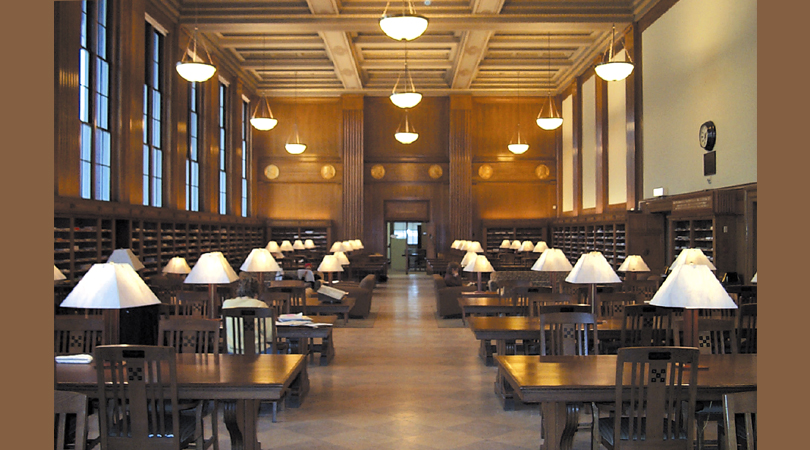 The Perodical Reading Room with long wooden tables, rows of bookshelves, elegant paneling, and table and ceiling lamps
