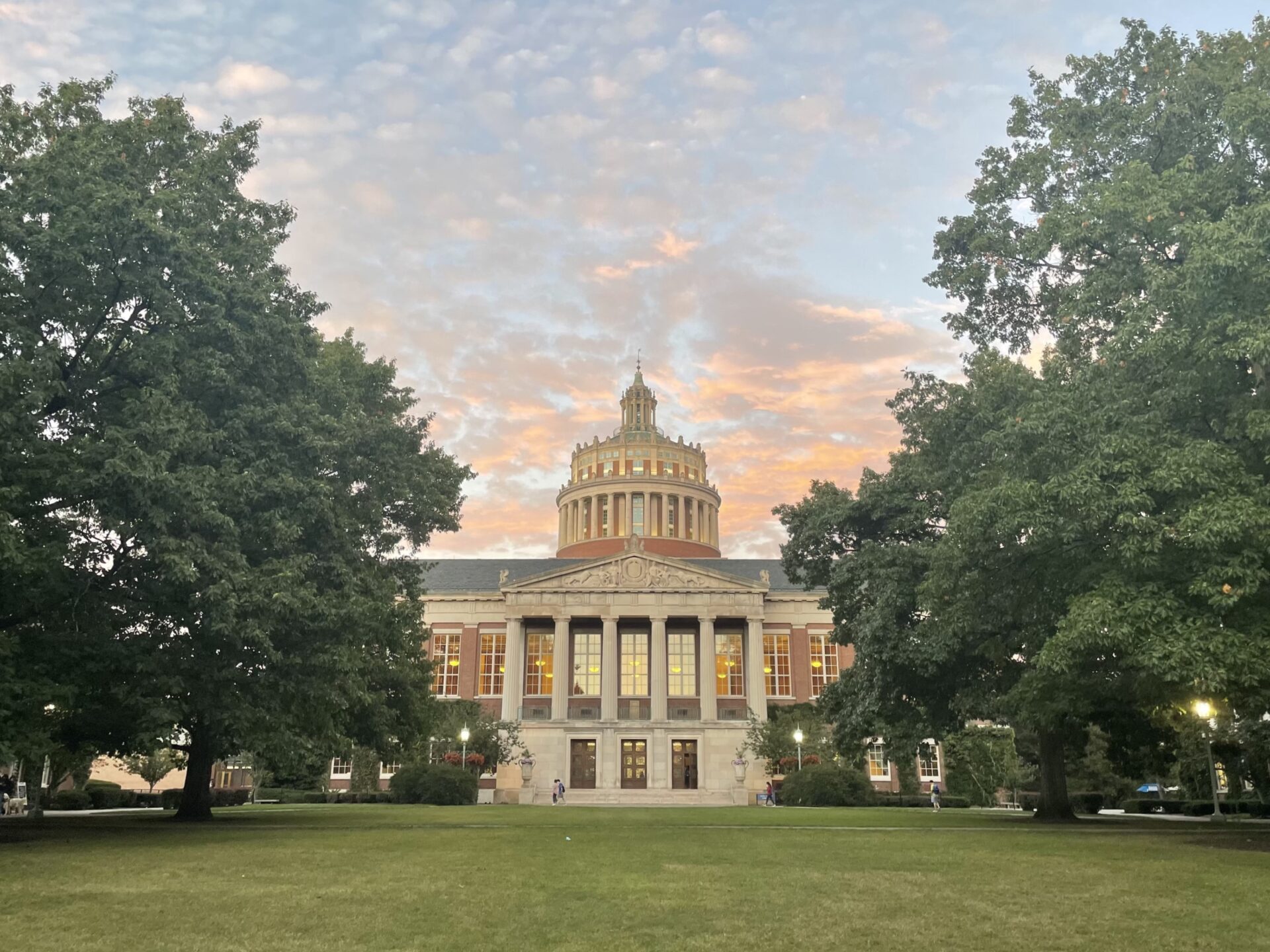 A pink, white, blue, and gray cloud sunset behind Rush Rhees on Eastman Quad flanked by big green trees