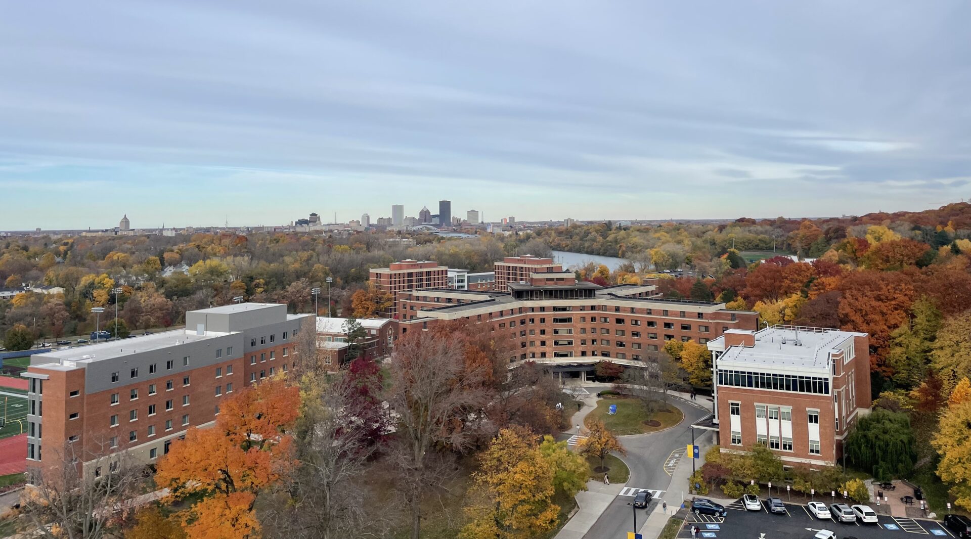 View of Genesee and Susan B Anthony Halls surrounded by orange fall foliage and city of Rochester skyline in the distance.