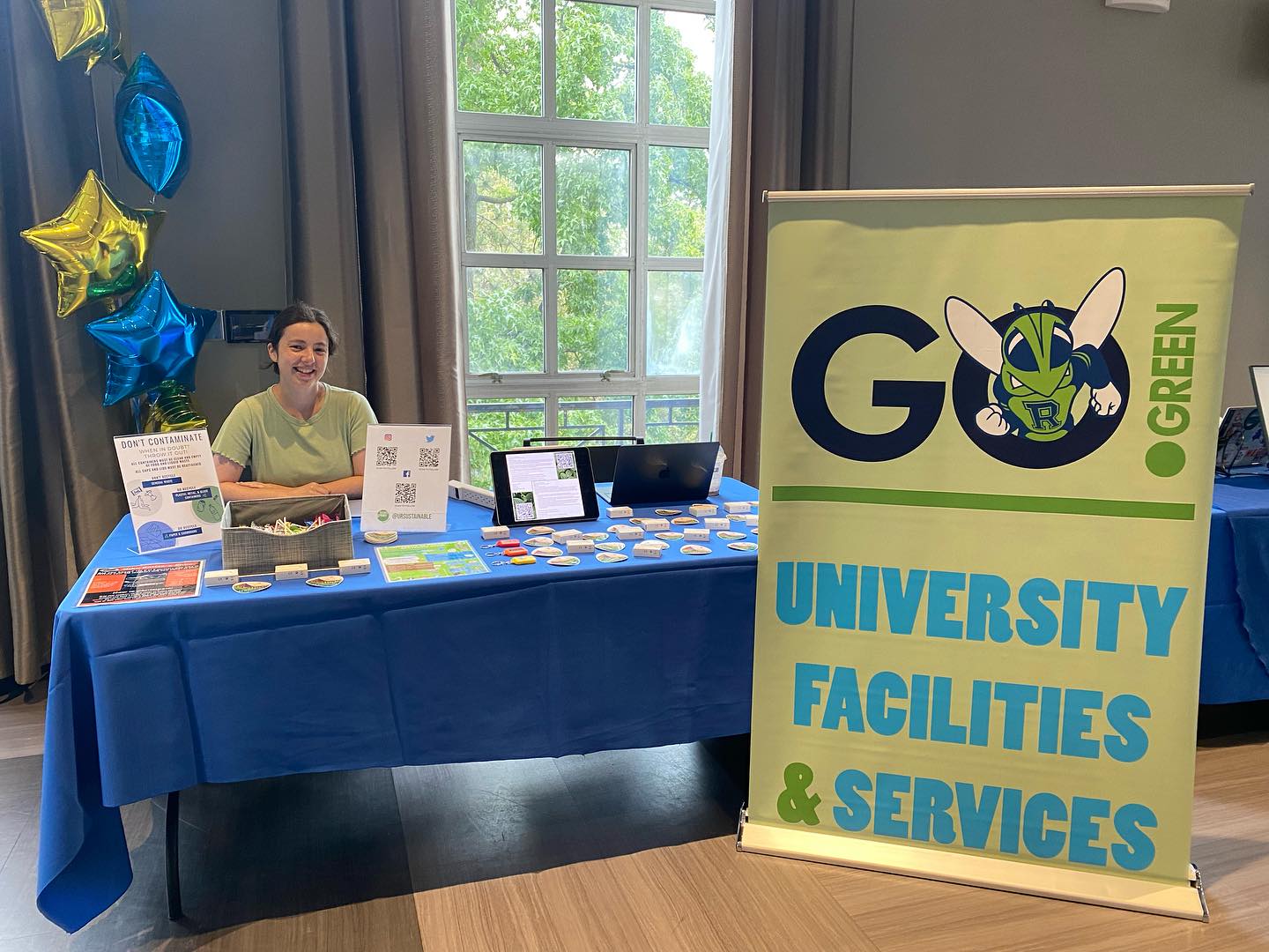 Girl in a green shirt sitting behind a table with a big sign saying Go Green University Facilities & Services