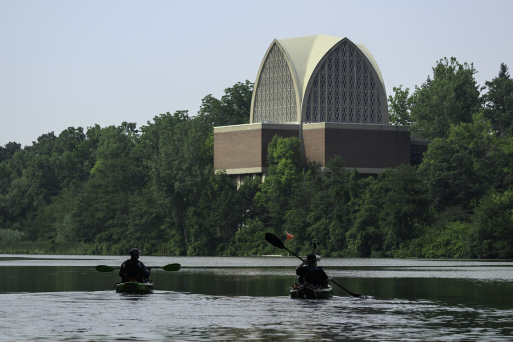 Two student kayak along the Genesee River, the Interfaith Chapel is seen in the background.