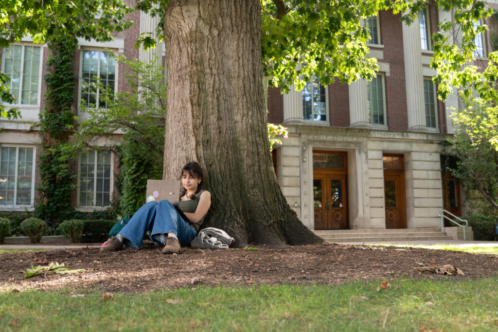 A student studies at the base of a large tree on Eastman Quad.