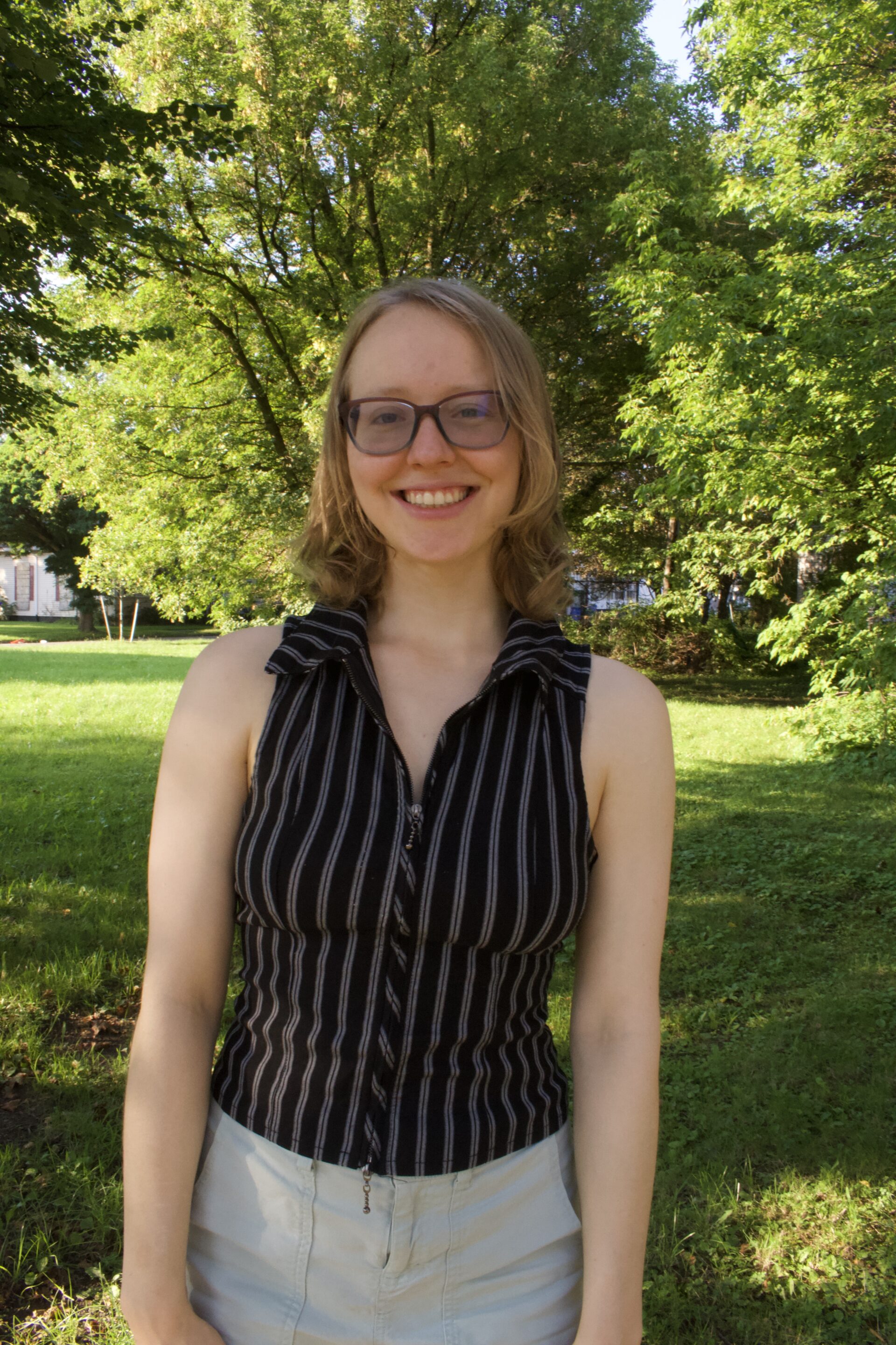 A URochester student stands in front of an area with many green trees. They are wearing a black blouse and have black glasses.