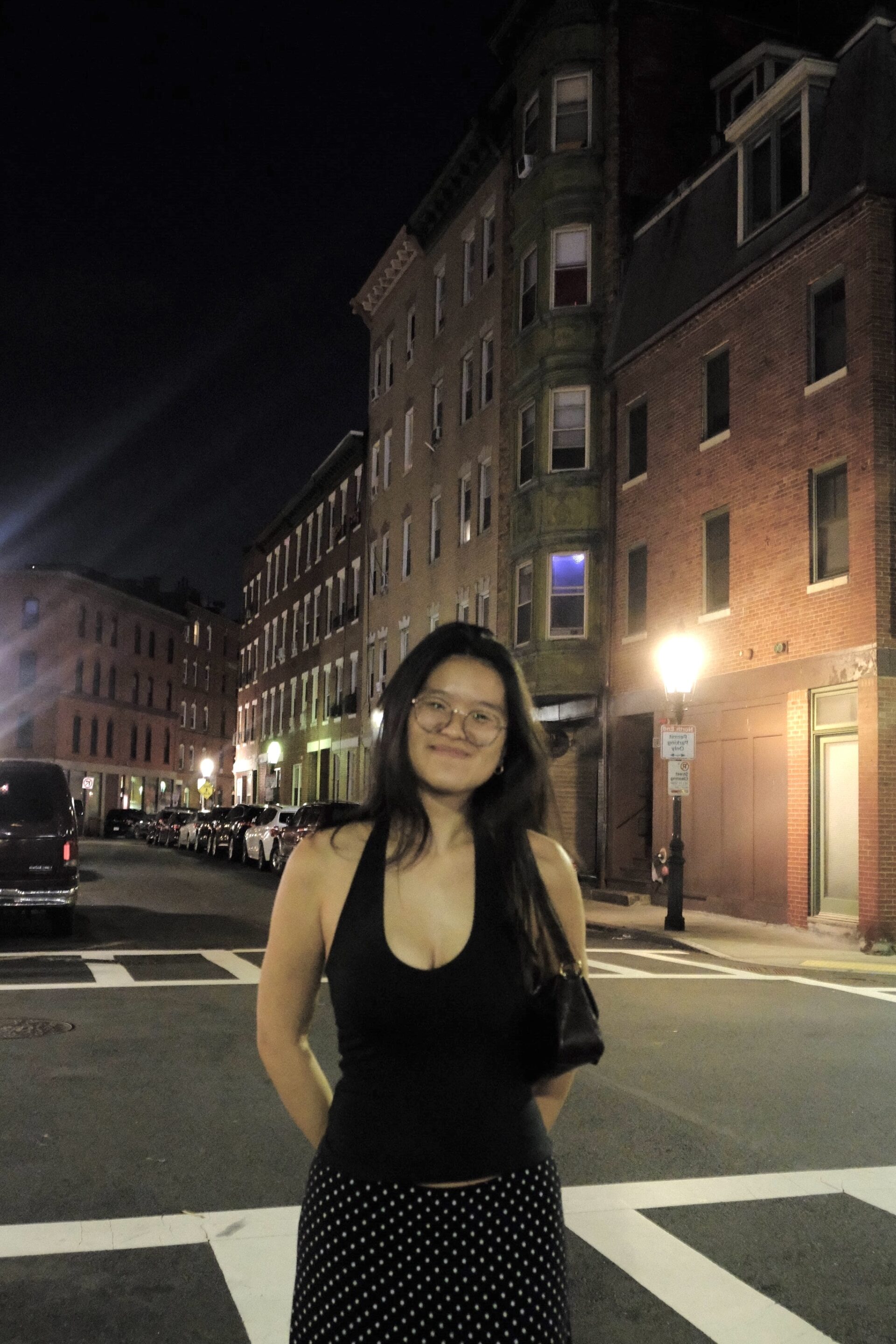 A student with long dark hair and round glasses stands in front of brick buildings. She is wearing a black halter neck shirt and a black and white polka dot skirt.