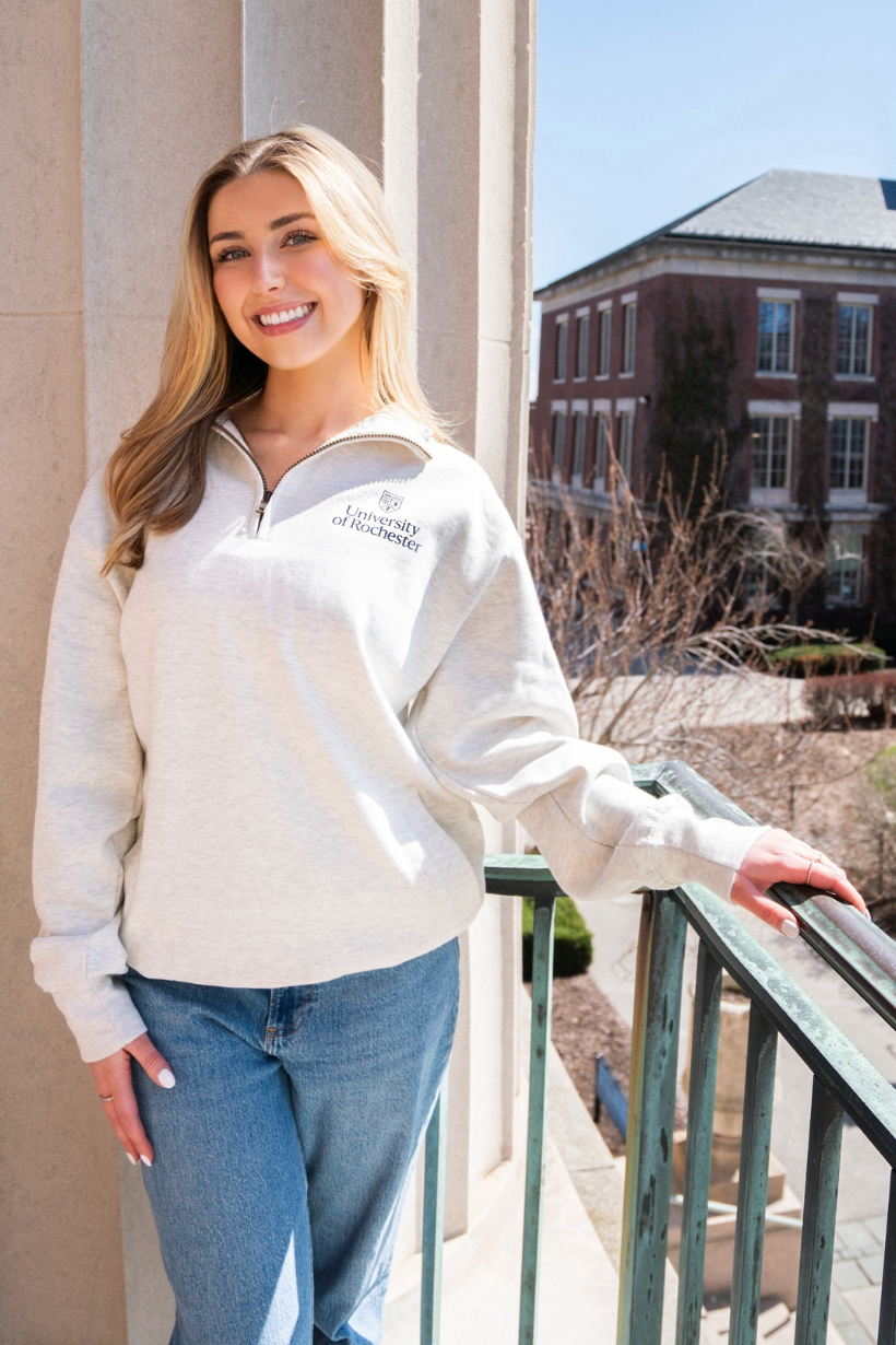 A student with long blonde hair stands in front of a pillar on the balcony of Rush Rhees Library. She wears a gray, URochester branded quarter zip sweatshirt and blue jeans.