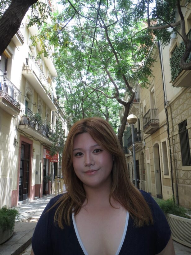 A student with long dark hair stands in front of white brick buildings and greenery. She is wearing a black shirt.