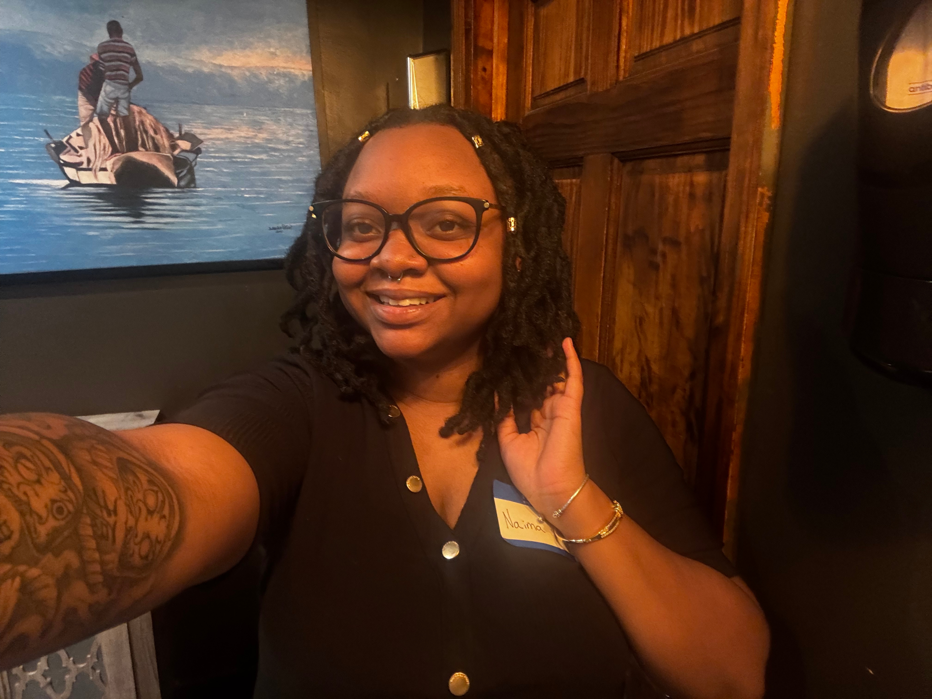 A URochester student sits in a room with a large blue painting behind them. They are wearing a black blouse and have black glasses.