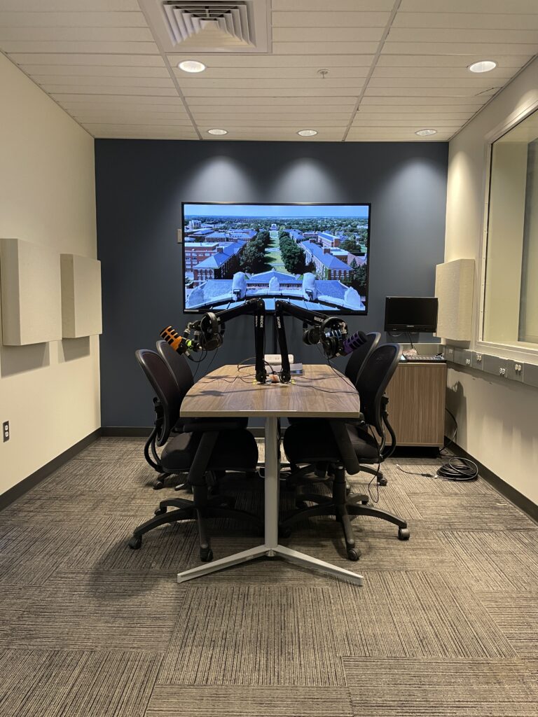 The podcast studio in Rettner Hall featuring a large recording device on a table in front of a large-screen TV. 