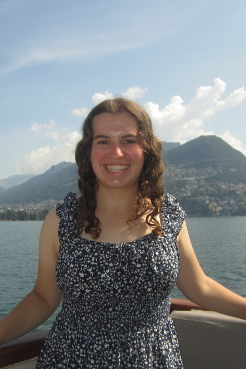 A student with long curly hair stands in front of scenic area with the ocean and a mountain in the distance. She is wearing a black, patterned dress.