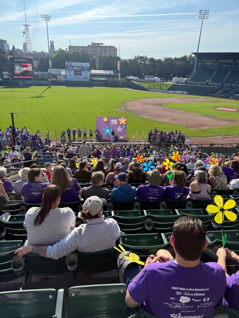 The backs of a crowd of people at a baseball stadium. They are sitting spread out in the seats and many wear the same purple shirt and hold flowers.