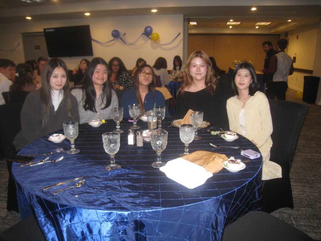 Five students sit around a round table that has a shiny, blue tablecloth at an event. 