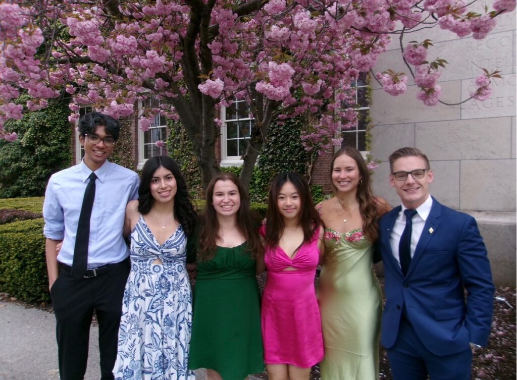 Six students in formal attire pose for a photo in front of a blooming cherry blossom tree on Eastman Quad. 
