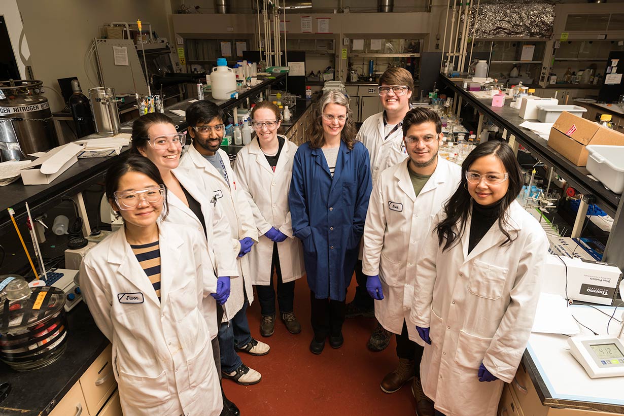 A group of students in labs coat with a professor all smiling at the camera.