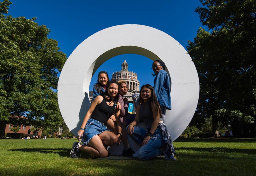A group of students posing with a large letter O with Rush Rhees Library in the background.