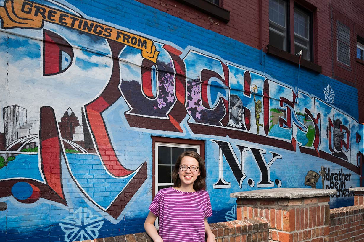 A young person standing in fron of a mural that says Greetings from Rochester, NY.