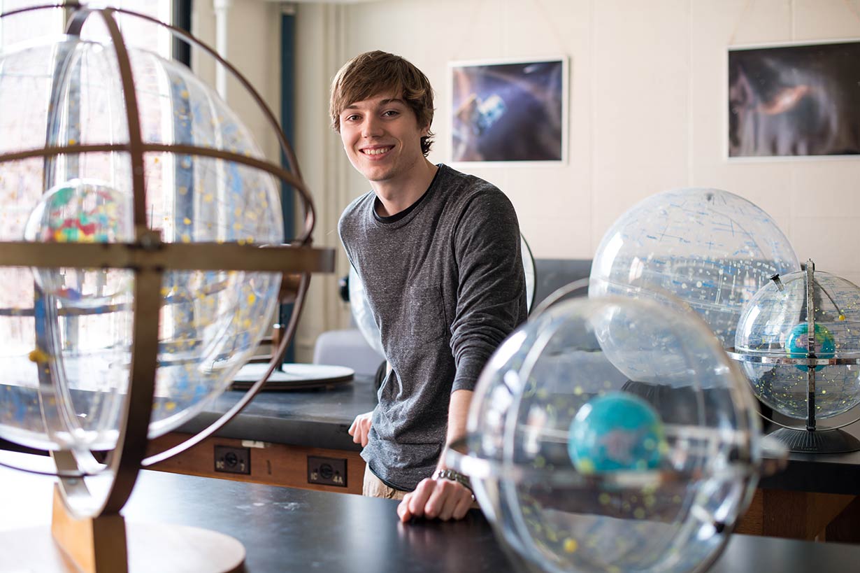 A senior student standing behind a desk surrounded by globes.
