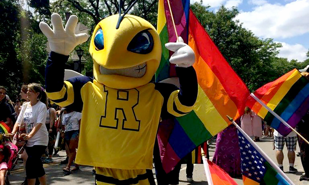 University of Rochester mascot, Rocky, the yellowjacket standing with a rainbow pride flag.