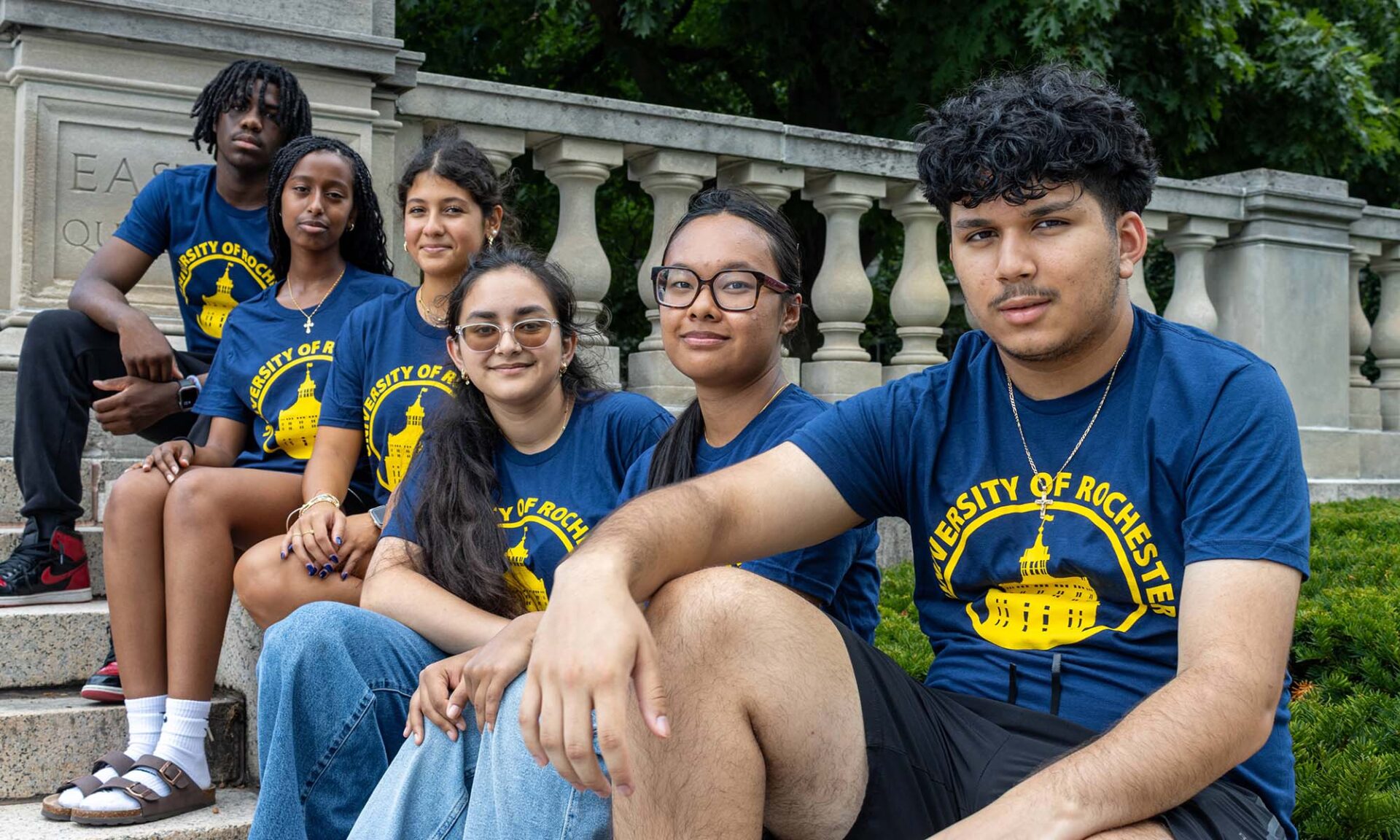 Half a dozen high school students wearing University of Rochester shirts sit on the stairs of the Eastman Quad as part of the University of Rochester's Pre-College Summer Program.