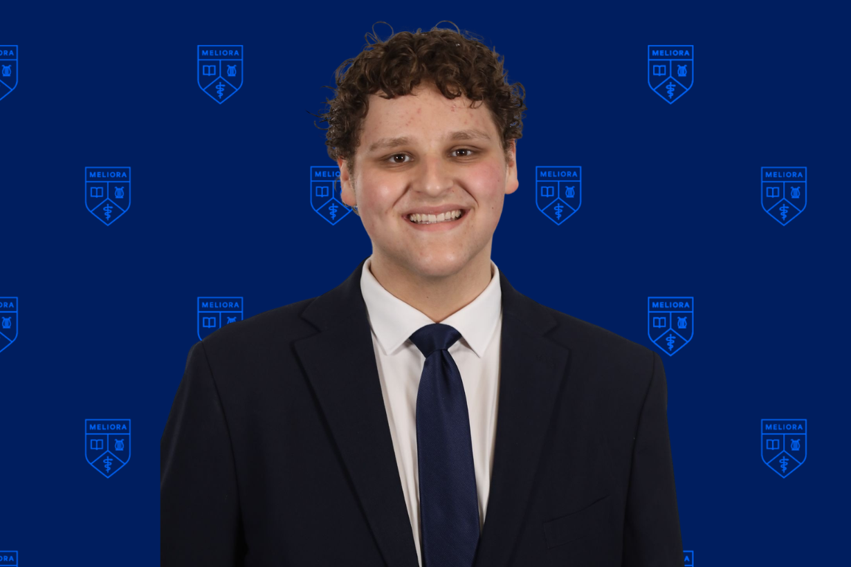 A URochester student in business attire in front of a navy background with a URochester shield pattern.