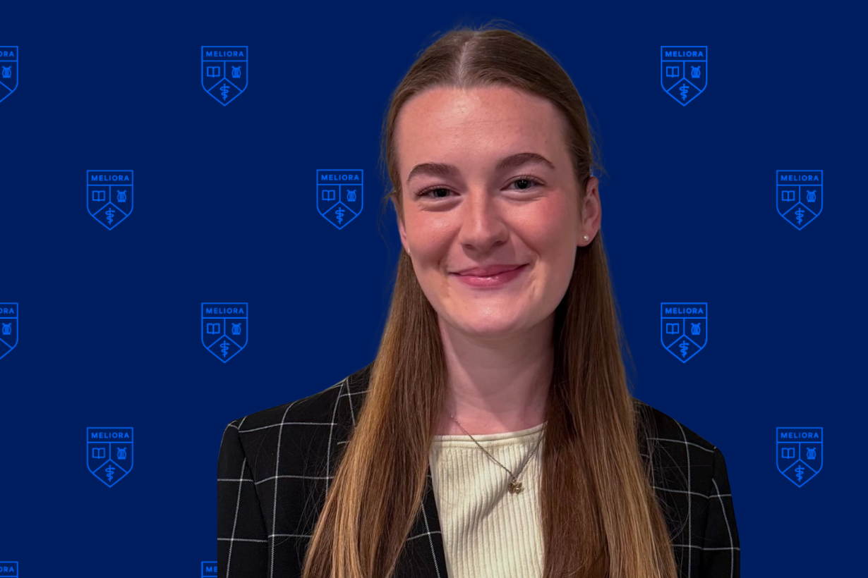A URochester student in a black patterned blazer in front of a navy background with a URochester shield pattern.