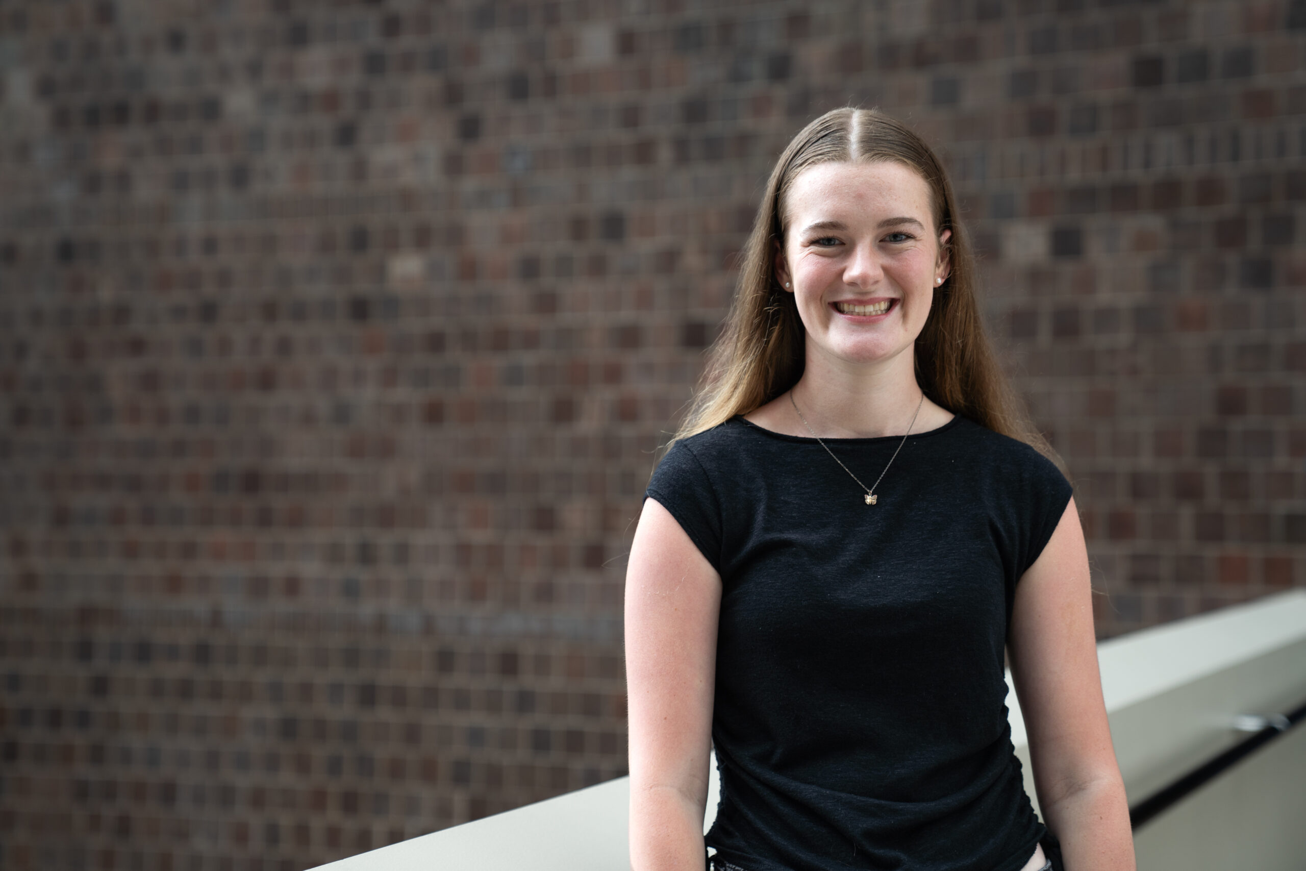 A URochester student with long hair, wearing a black shirt in front of a brick background.