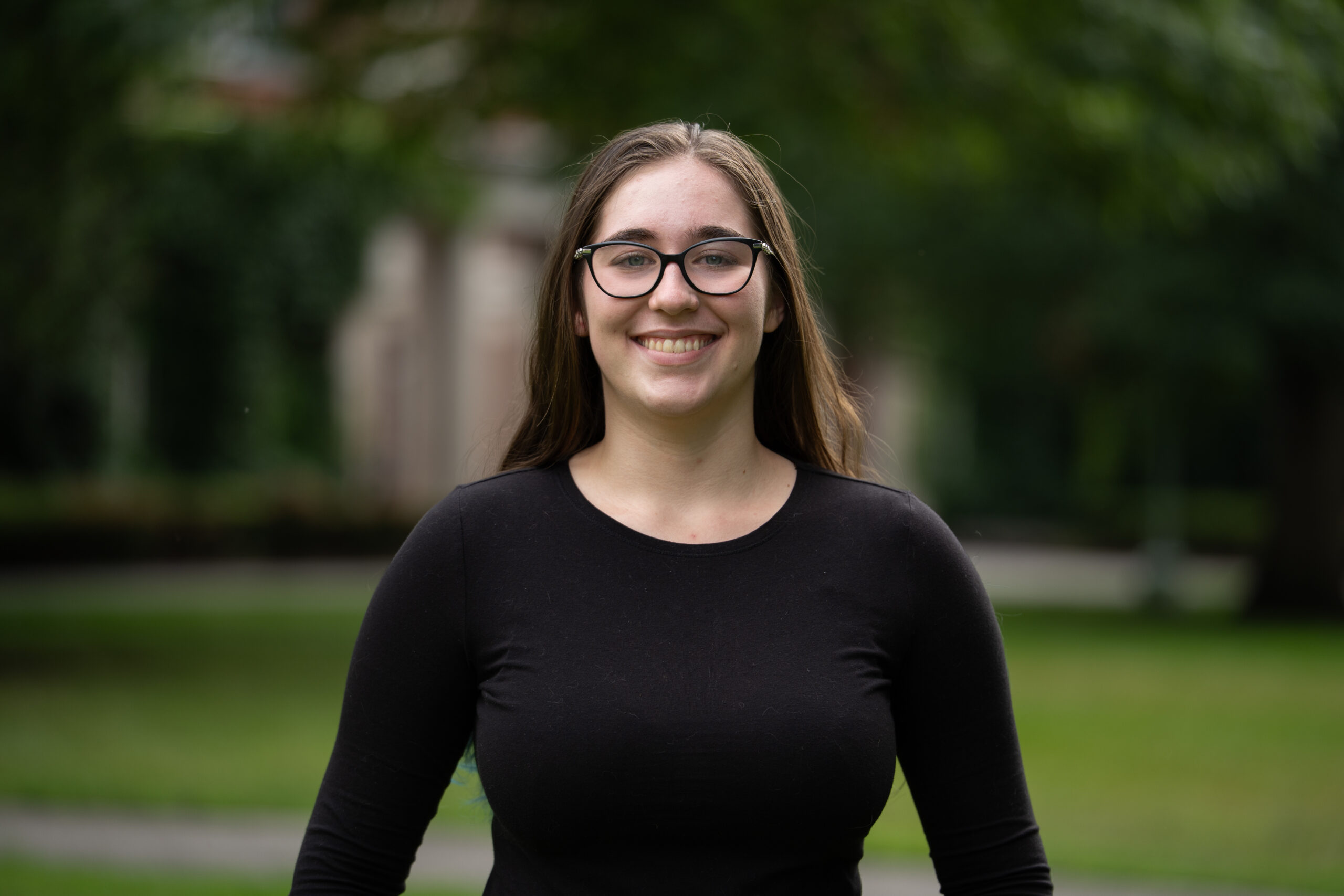 A student with long dark hair and glasses wears a long-sleeved black t-shirt in front of a greenery-filled area on Eastman Quad.