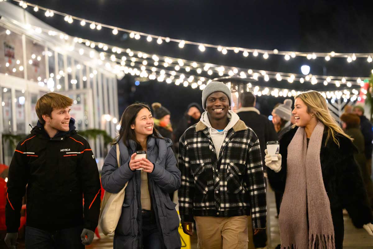 Four students walk and talk beneath strings of lights on a winter evening.