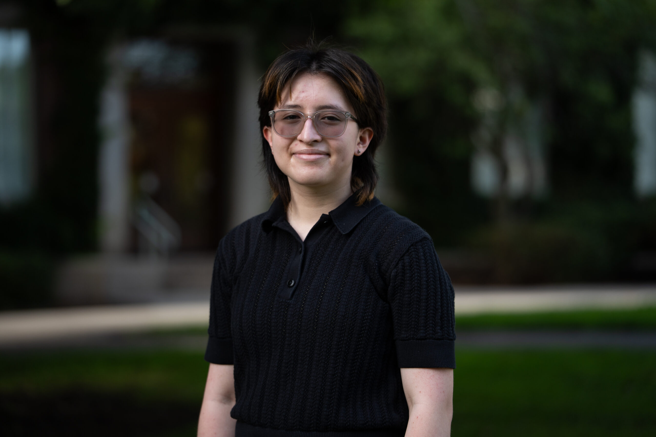 A student with short, dark brown hair wears a black, knit polo shirt on Eastman Quad.
