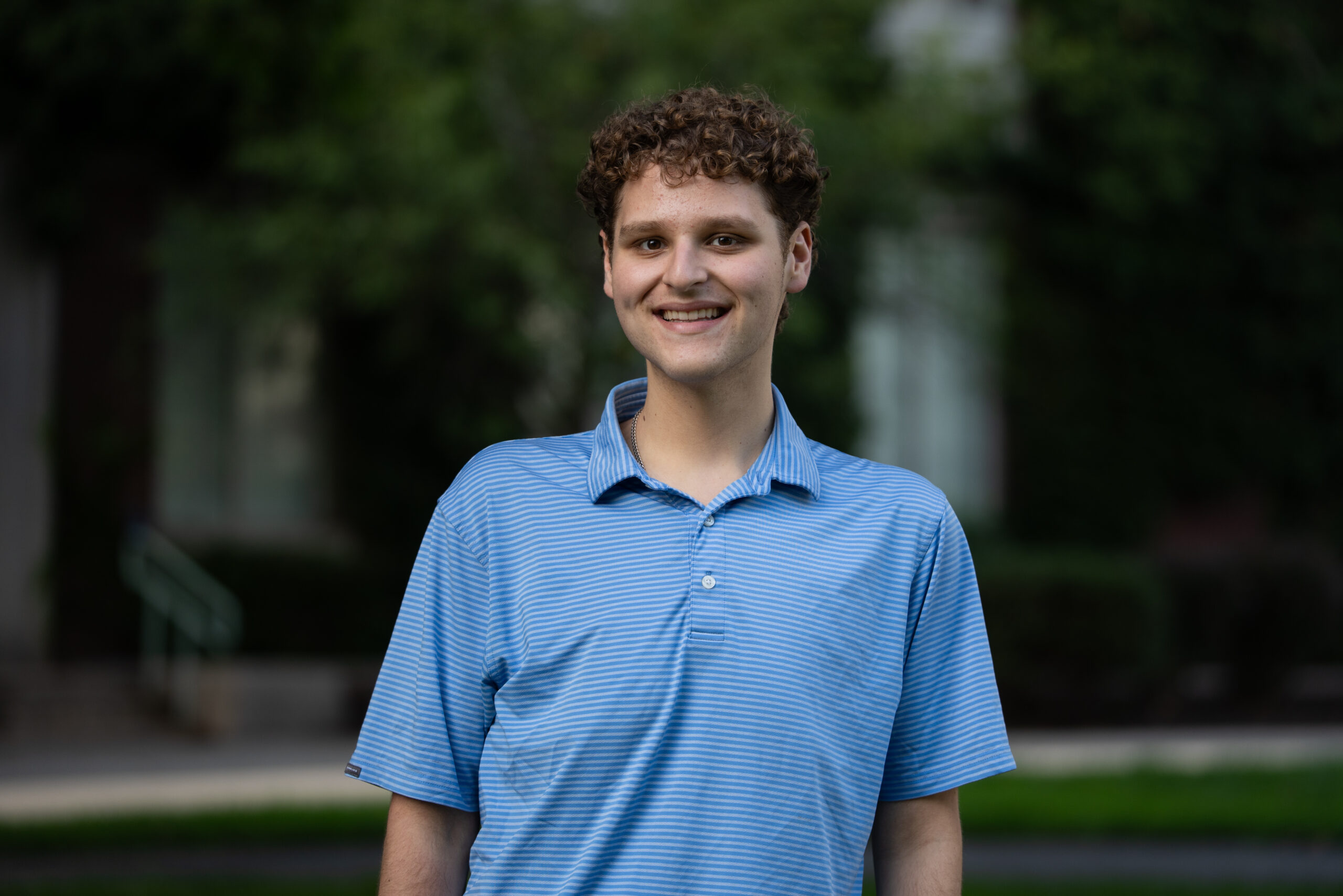 A student with short, curly brown hair wears a blue polo shirt on Eastman Quad.