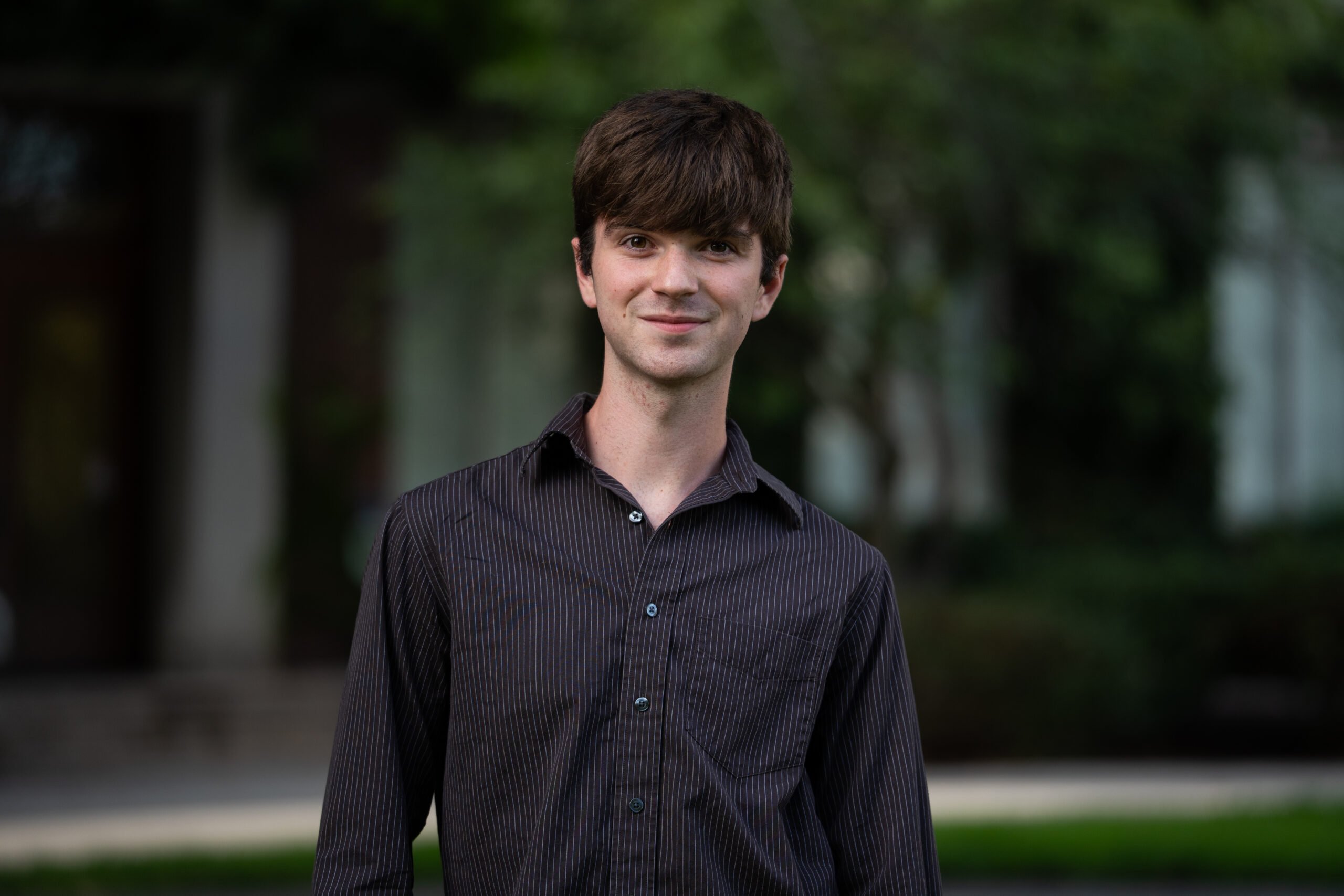 A student with short dark hair wears a long-sleeved dark, striped dress shirt in front of a greenery-filled area on Eastman Quad.