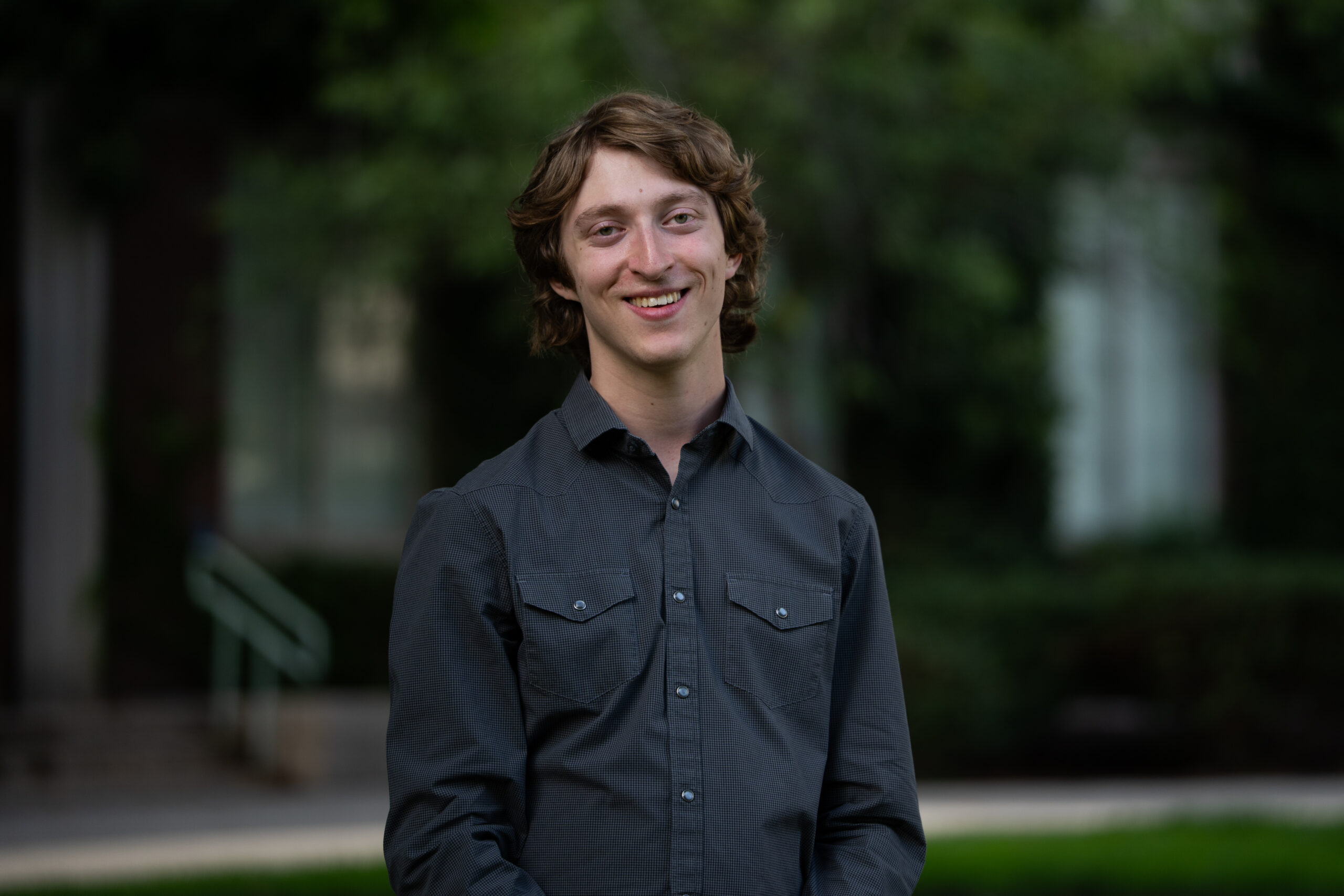 A student with wavy, brown hair wears a dark gray button-up shirt on Eastman Quad.