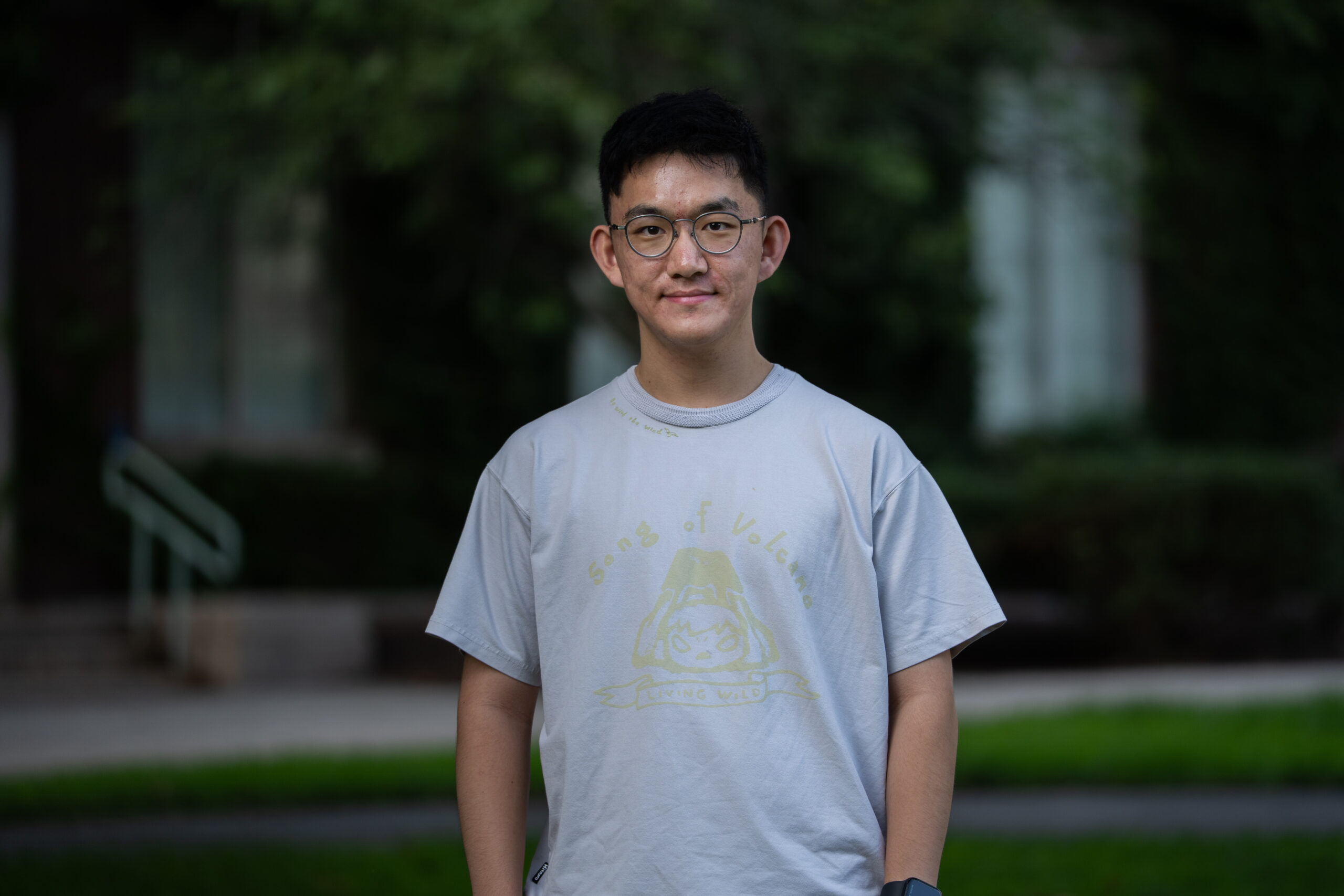 A student with short, black hair and glasses wears a light gray graphic t-shirt on Eastman Quad.