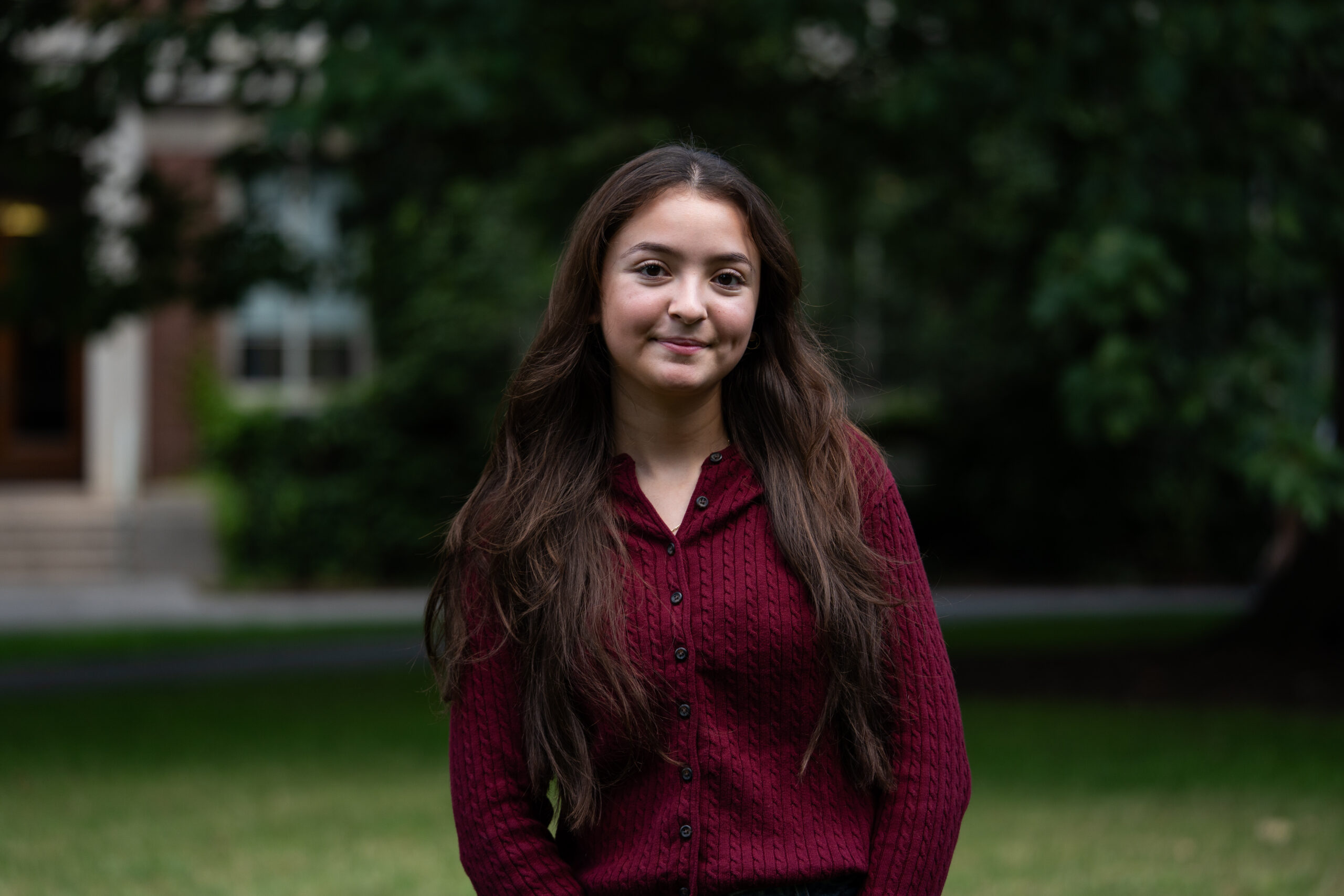 A student with long dark hair wears a long-sleeved maroon blouse in front of a greenery-filled area on Eastman Quad.