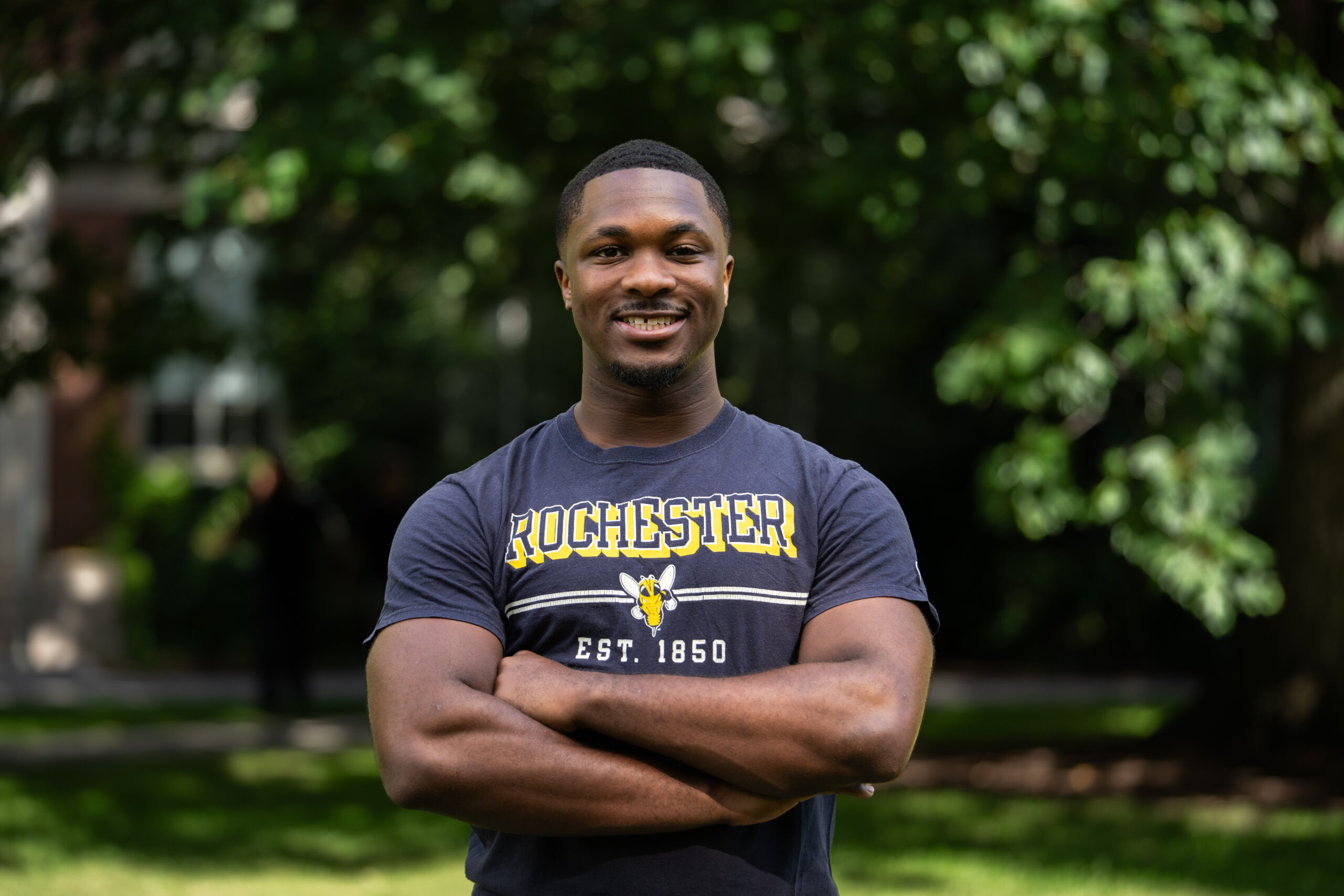 A student stands in "Rochester" t-shirt on on Eastman Quad with his arms crossed.