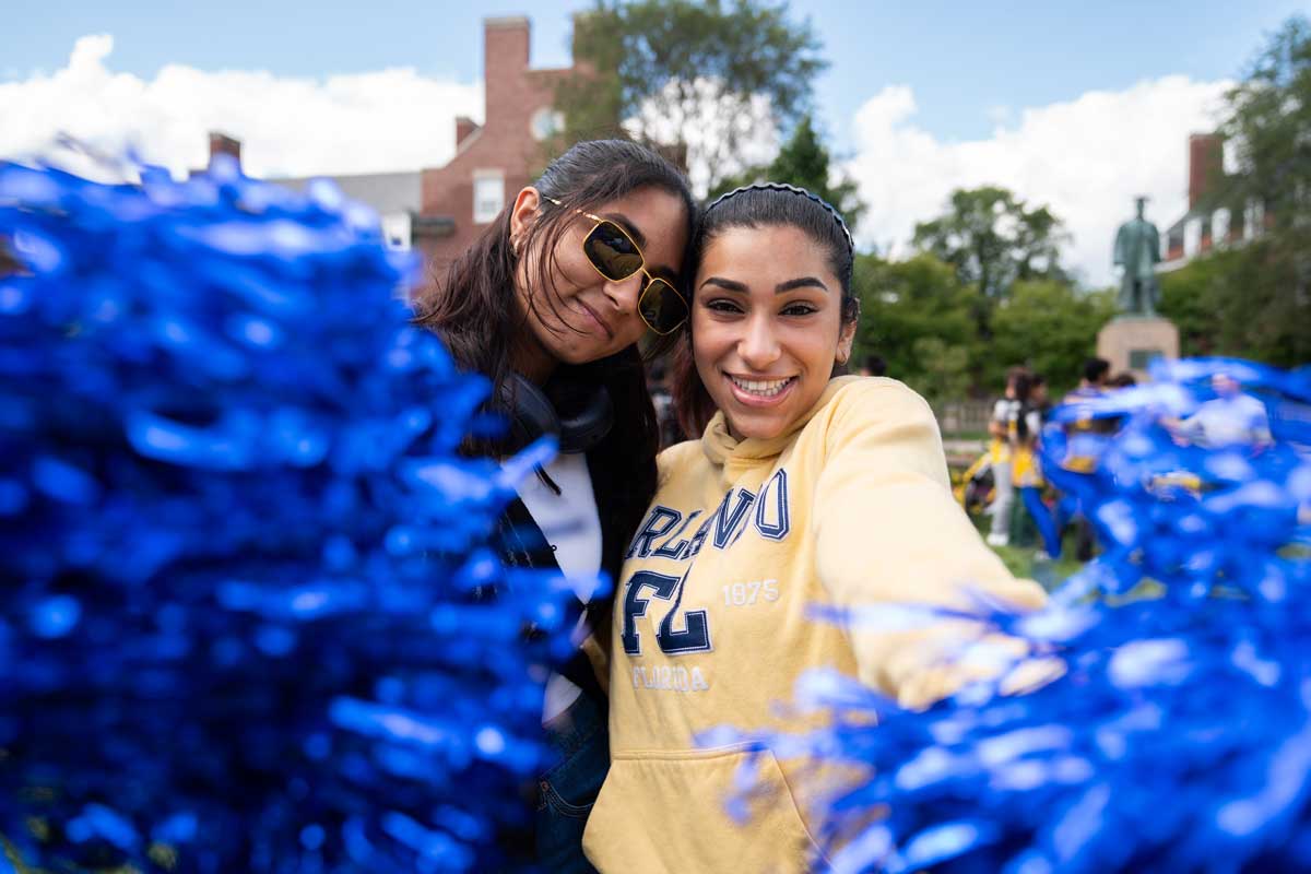 Two students smile behind bright blue pom-poms at an outdoor activities fair.