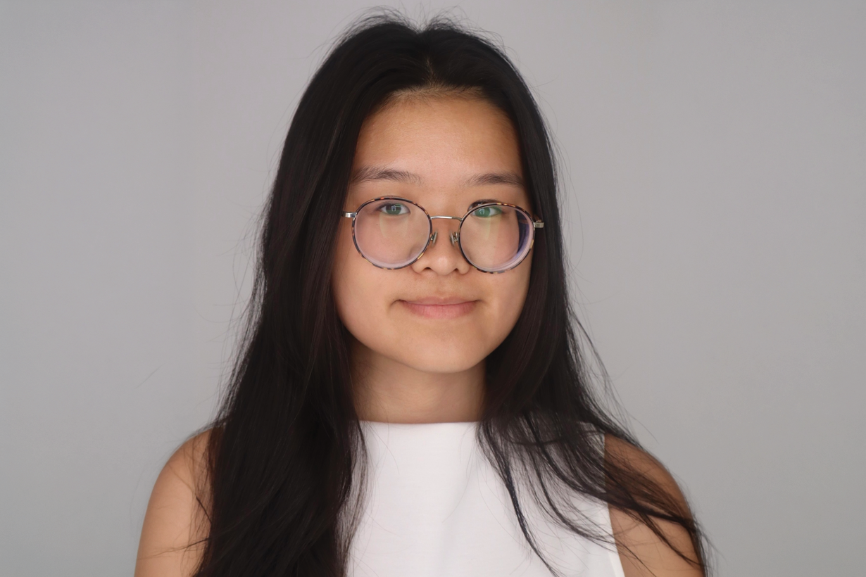 A student in a white, sleeveless blouse in front of a gray backdrop. She has long dark hair and round glasses.