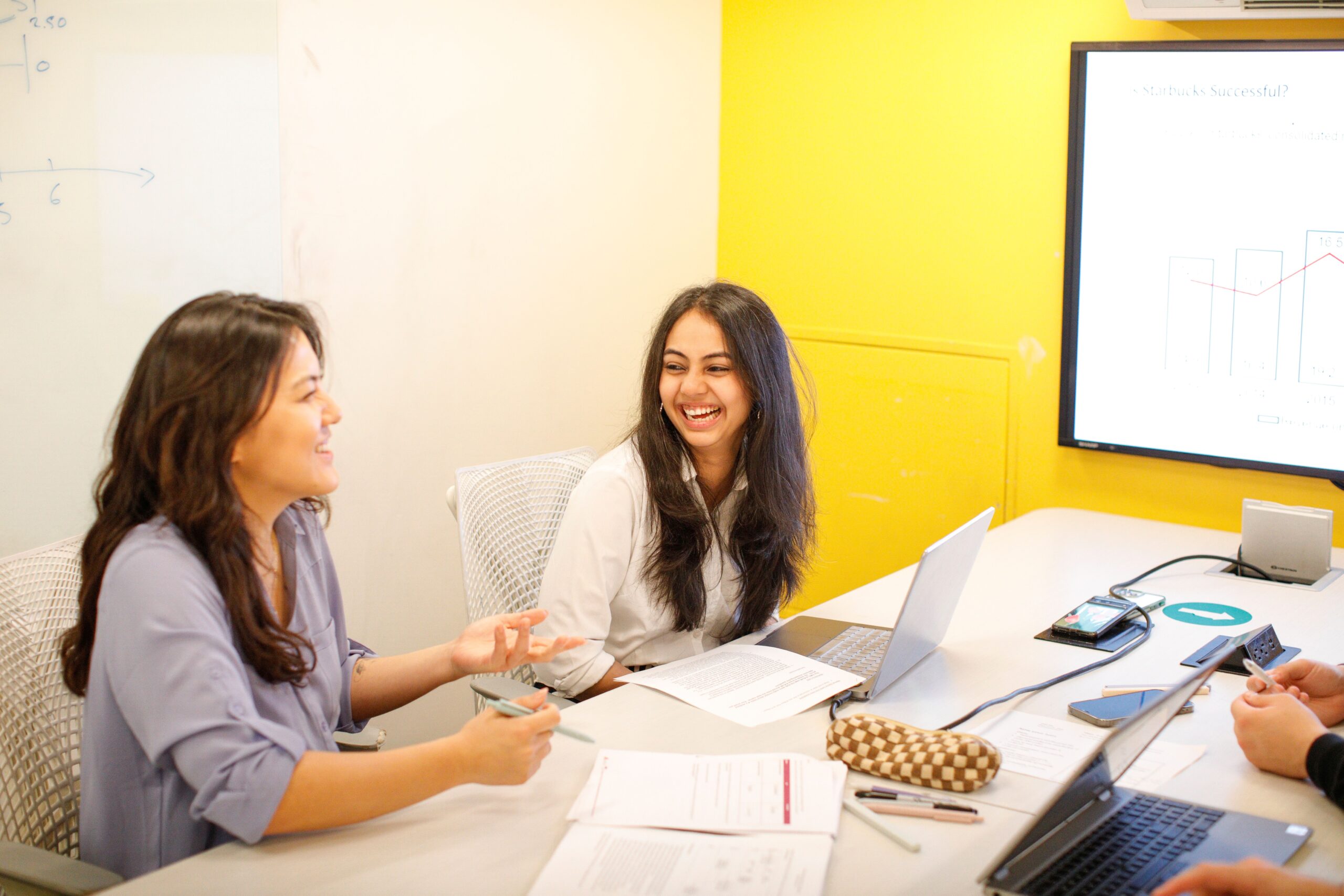 Two students chat at a table while working on project in front of a bright yellow wall.